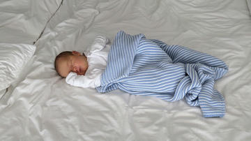 Photo of a sleeping baby boy wearing a white onesie and wrapped in a blue blanket, laying on a big white duvet on his parent's bed.